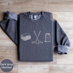 May include: Dark gray heather sweatshirt with white line art of a sandwich, crossed hockey sticks, a hockey puck, and a ginger ale can. The text "Dark Gray Heather" is at the bottom left.