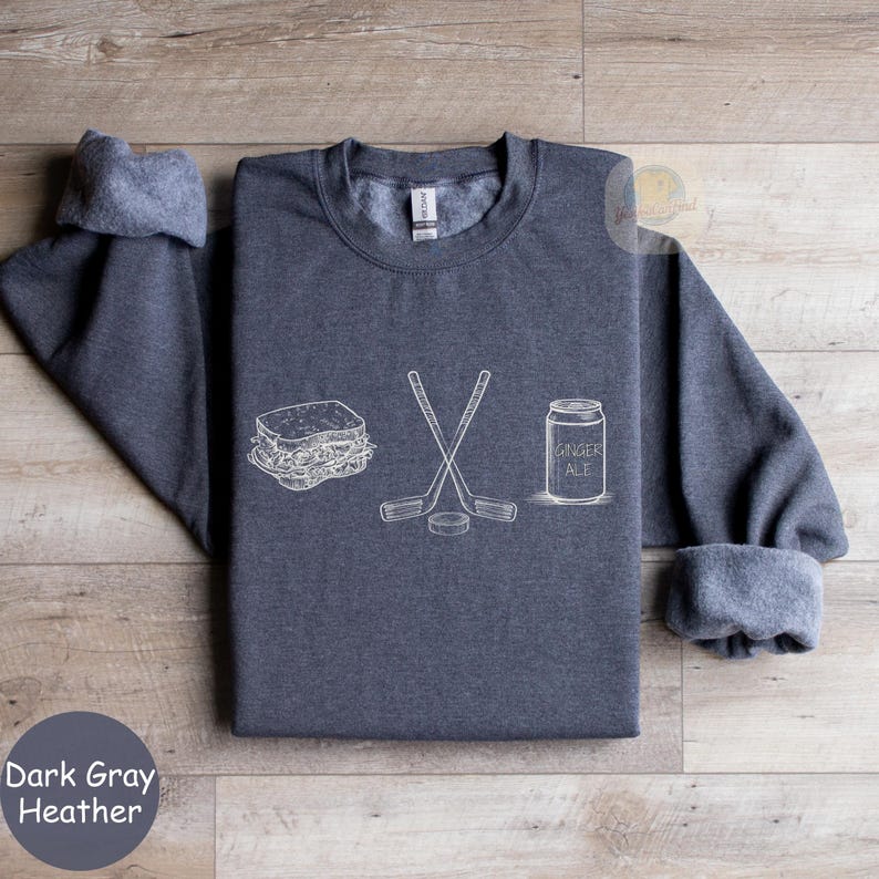 May include: Dark gray heather sweatshirt with white line art of a sandwich, crossed hockey sticks, a hockey puck, and a ginger ale can. The text "Dark Gray Heather" is at the bottom left.