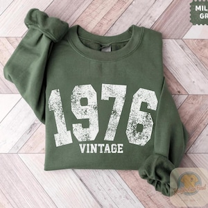 Può includere: Una felpa verde militare con l'anno "1976" stampato in grandi lettere bianche usurate. La parola "VINTAGE" è stampata sotto. Le maniche sono arrotolate. La felpa è esposta su una superficie di legno chiaro.