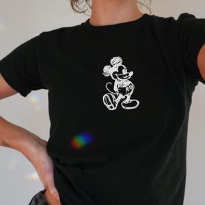 May include: Black t-shirt featuring a white sketch of Mickey Mouse. The short-sleeved shirt has a crew neck and is made of a soft material. The design is a simple, line-art style illustration of the iconic cartoon character.