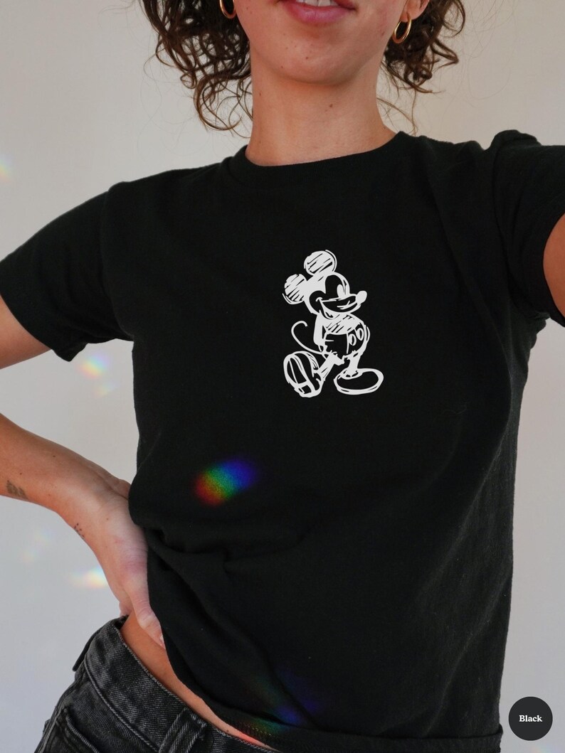 May include: Black t-shirt featuring a white sketch of Mickey Mouse. The short-sleeved shirt has a crew neck and is made of a soft material. The design is a simple, line-art style illustration of the iconic cartoon character.