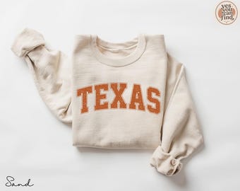 Könnte beinhalten: Ein sandfarbenes Sweatshirt mit Rundhalsausschnitt und dem Wort TEXAS, das in einer abgenutzten, rostorangen College-Schrift auf der Brust gedruckt ist. Das Kleidungsstück hat lange Ärmel mit gerippten Bündchen und einen gerippten Halsausschnitt.