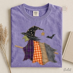 May include: A violet t-shirt featuring a graphic of a witch on a broomstick. The witch wears a black hat with stars and a moon, and a cloak made of patchwork fabric in purple plaid, orange gingham, and gray stripes. A small yellow star and a black bat are nearby.