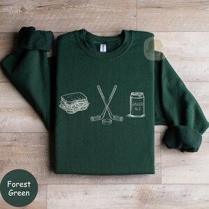 May include: Forest green sweatshirt with a white graphic of a sandwich, crossed hockey sticks, a hockey puck, and a ginger ale can. The sweatshirt has a crew neck and long sleeves.