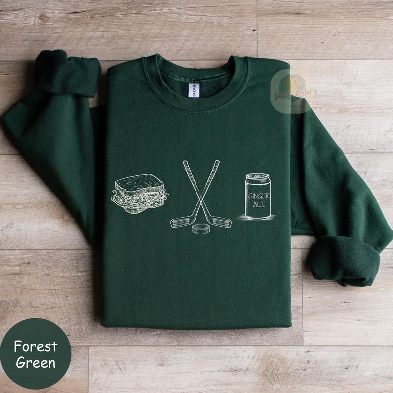 May include: Forest green sweatshirt with a white graphic of a sandwich, crossed hockey sticks, a hockey puck, and a ginger ale can. The sweatshirt has a crew neck and long sleeves.