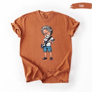 May include: A yam-colored t-shirt featuring a cartoon illustration of a musician playing a guitar. The illustration depicts a man with gray hair, a white beard, a white shirt, blue shorts, and black sandals. The shirt has rolled-up sleeves.