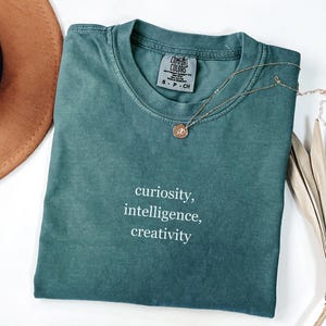 Puede incluir: Una camiseta Comfort Colors verde azulado con las palabras "curiosity, intelligence, creativity" impresas en blanco. Un collar dorado con un pequeño colgante descansa sobre la camiseta. Un sombrero marrón y una hoja de palma seca están en el fondo.