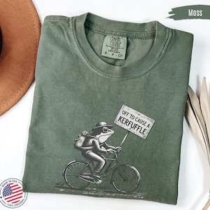 May include: A moss green t-shirt featuring a frog on a bicycle, holding a sign saying "OFF TO CAUSE A KERFUFFLE." The shirt has a crew neck and short sleeves. The t-shirt is made in the USA.