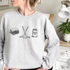 May include: Sport gray sweatshirt with a black and white graphic of a sandwich, crossed hockey sticks, a hockey puck, and a ginger ale can. The text "GINGER ALE" is on the can.