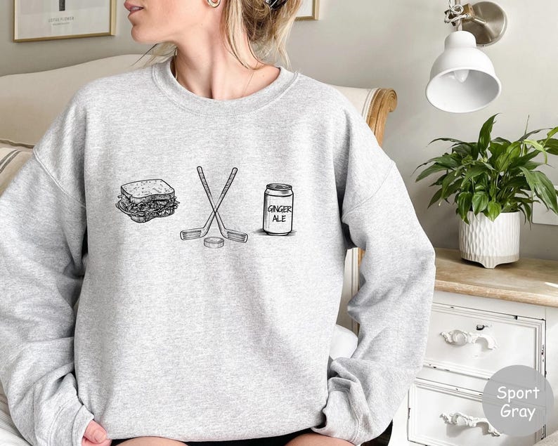 May include: Sport gray sweatshirt with a black and white graphic of a sandwich, crossed hockey sticks, a hockey puck, and a ginger ale can. The text "GINGER ALE" is on the can.