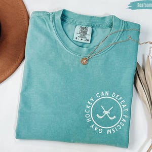 May include: Seafoam green t-shirt with the text "GAY HOCKEY CAN DEFEAT FASCISM" in a circular design. The shirt has a crew neck and a small tag on the upper chest. A gold necklace with a pendant is draped across the shirt.