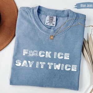 Puede incluir: Camiseta azul vaquero con el texto "F**CK ICE SAY IT TWICE" en letras blancas. Un collar dorado con un colgante está drapeado sobre la camisa. La camisa está hecha de un material suave.