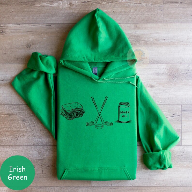 May include: Irish green hoodie with a hood and long sleeves. The front features a black line drawing of a sandwich, crossed hockey sticks, a hockey puck, and a can of ginger ale. The text "GINGER ALE" is on the can.