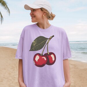 May include: A light purple t-shirt featuring a graphic of two red cherries with a green leaf. The cherries have a glossy appearance. The shirt is worn by a person wearing a white cap and gold earrings.
