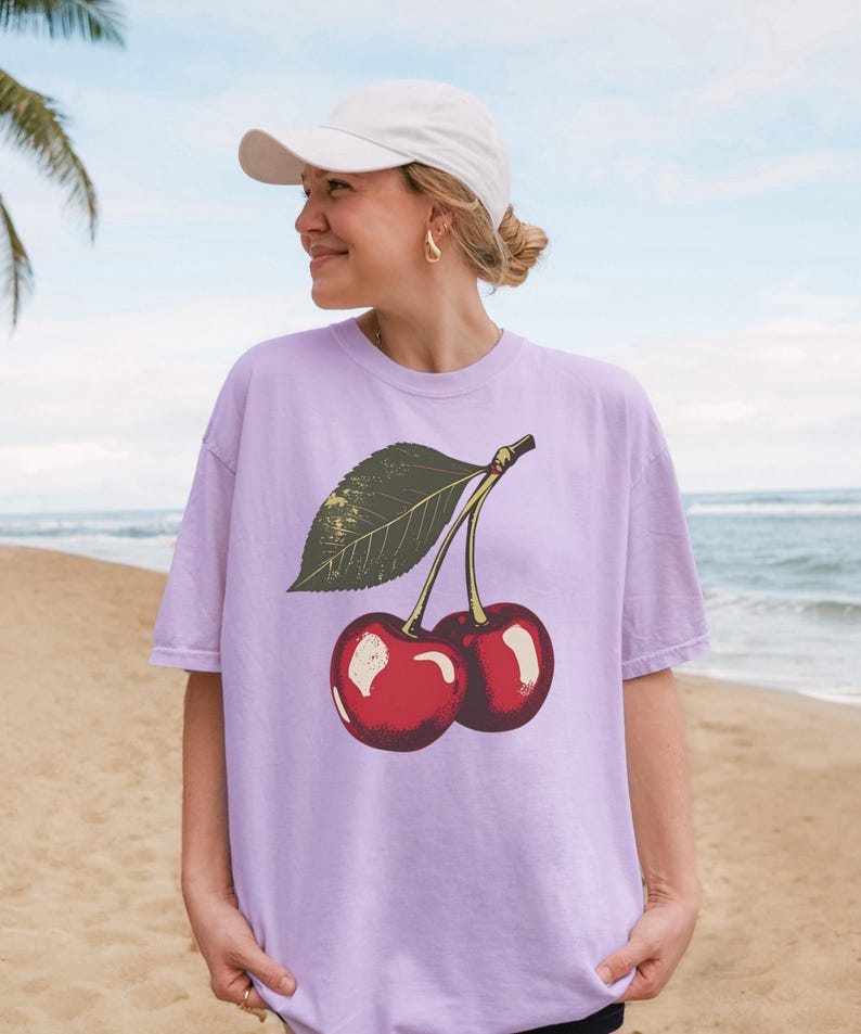 May include: A light purple t-shirt featuring a graphic of two red cherries with a green leaf. The cherries have a glossy appearance. The shirt is worn by a person wearing a white cap and gold earrings.