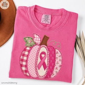 May include: A bright pink short-sleeved t-shirt featuring a large pumpkin applique on the front. The pumpkin is constructed from various fabric patches in pink and green, including gingham, polka dots, and floral patterns, with a pink ribbon.