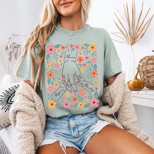 May include: A light green t-shirt featuring a floral design with the words "Nana", "Ryan", and "Emily". The graphic shows hands holding each other, surrounded by colorful flowers. The shirt is made of a soft material.
