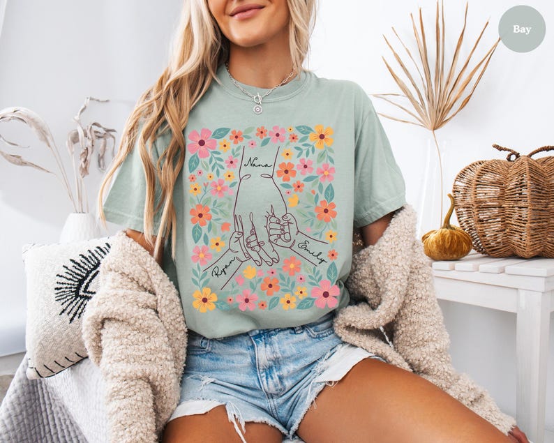 May include: A light green t-shirt featuring a floral design with the words "Nana", "Ryan", and "Emily". The graphic shows hands holding each other, surrounded by colorful flowers. The shirt is made of a soft material.
