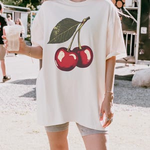 May include: Cream-colored oversized t-shirt featuring a vintage-style illustration of two ripe cherries with a green leaf. The cherries are a vibrant red with white highlights. The shirt has short sleeves and a relaxed fit, suitable for casual wear.
