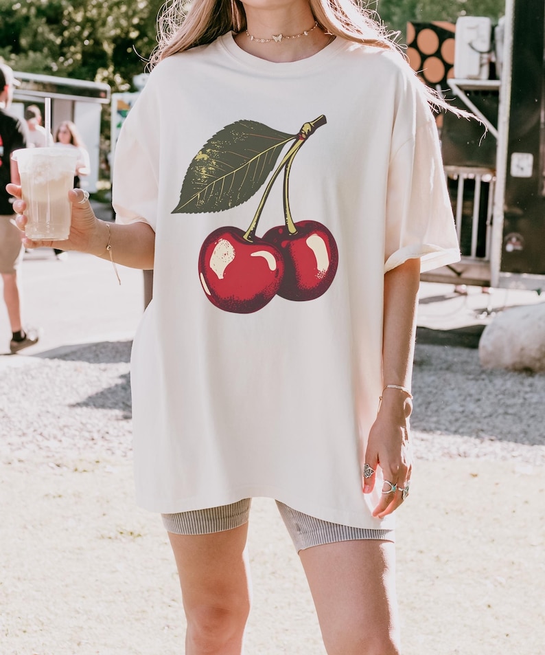May include: Cream-colored oversized t-shirt featuring a vintage-style illustration of two ripe cherries with a green leaf. The cherries are a vibrant red with white highlights. The shirt has short sleeves and a relaxed fit, suitable for casual wear.