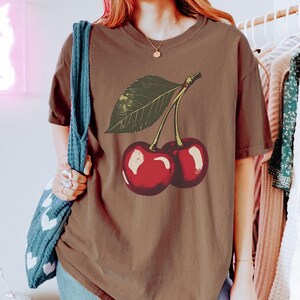 May include: A brown t-shirt featuring a graphic of two cherries with a green leaf. The cherries are red with white highlights. The shirt has short sleeves and a crew neck. The person is wearing blue jeans and a blue tote bag.
