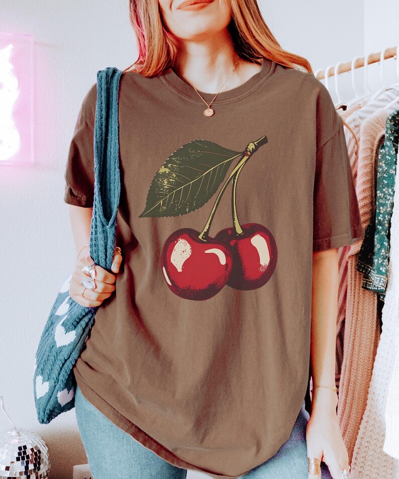 May include: A brown t-shirt featuring a graphic of two cherries with a green leaf. The cherries are red with white highlights. The shirt has short sleeves and a crew neck. The person is wearing blue jeans and a blue tote bag.