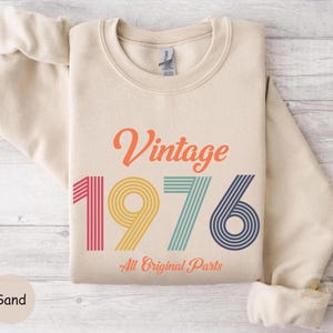 May include: A sand-colored sweatshirt with the text "Vintage 1976 All Original Parts" in orange, pink, yellow, teal, and navy blue. The sweatshirt is folded, with the sleeves rolled up. The background is a light wood grain.