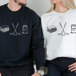 May include: Black and white graphic design on a black sweatshirt and a white sweatshirt. The design includes a sandwich, crossed hockey sticks, a hockey puck, and a can of ginger ale. The text "GINGER ALE" is on the can.