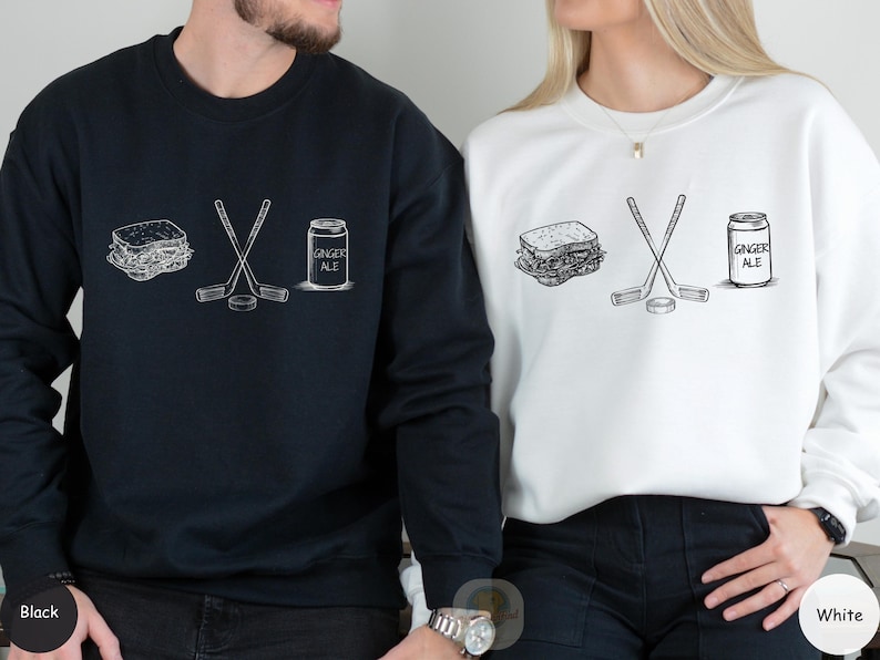 May include: Black and white graphic design on a black sweatshirt and a white sweatshirt. The design includes a sandwich, crossed hockey sticks, a hockey puck, and a can of ginger ale. The text "GINGER ALE" is on the can.