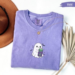 Może przedstawiać: Fioletowy t-shirt z krótkim rękawem z wyhaftowaną grafiką białego ducha trzymającego fioletową puszkę Alani. Koszulka ma okrągły dekolt i metkę marki Comfort Colors wewnątrz kołnierza. Wokół ducha znajdują się małe czarne nietoperze i gwiazdki.