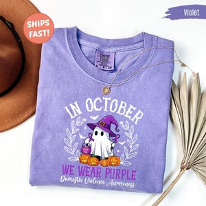 May include: A violet short-sleeved t-shirt featuring a graphic of a ghost wearing a purple witch hat, holding a small pumpkin bucket, and surrounded by pumpkins. The text reads: In October we wear purple, Domestic Violence Awareness.