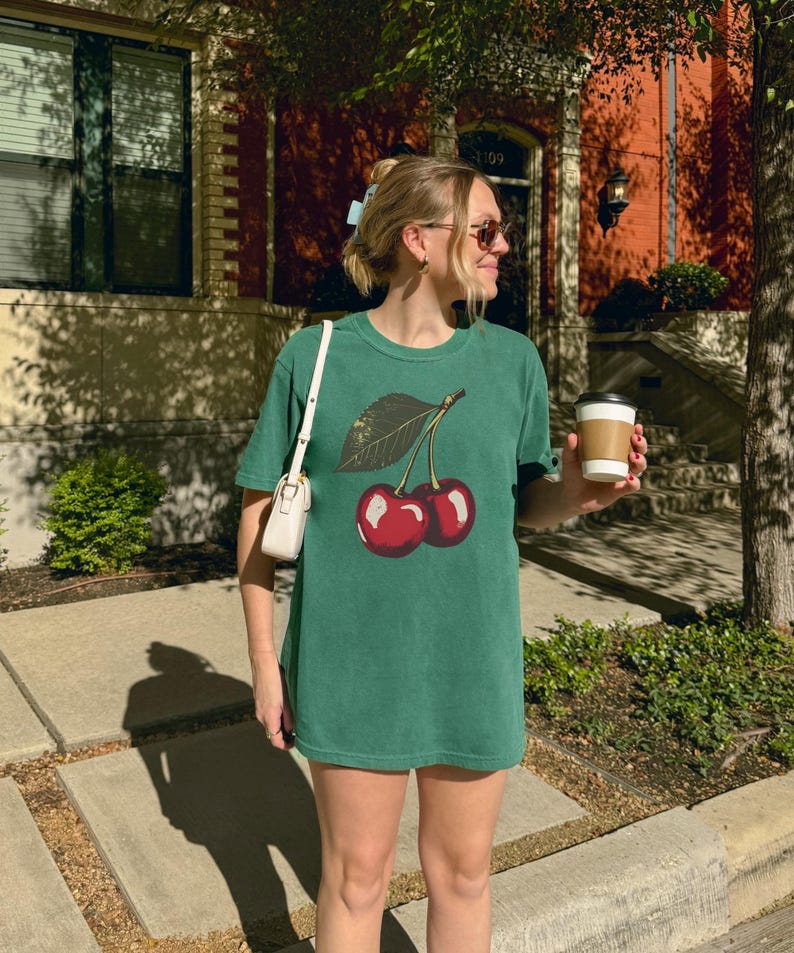 May include: Green t-shirt featuring a graphic of two red cherries with a green leaf. The shirt is a solid green color and appears to be made of a soft material. The person is wearing sunglasses and carrying a white purse and a coffee cup.