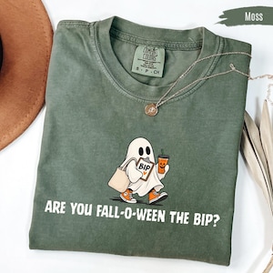 Könnte beinhalten: Ein moosgrünes Kurzarm-T-Shirt mit einer Grafik eines weißen Geistes, der eine Tragetasche, ein Klemmbrett mit der Aufschrift BIP und einem Herz sowie ein orangefarbenes Getränk trägt. Der Text lautet ARE YOU FALL-O-WEEN THE BIP? Comfort Colors Etikett.