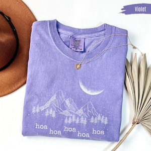 May include: A violet Comfort Colors t-shirt with a mountain and moon graphic. The design includes trees and the word "hoa" repeated. A gold necklace and a brown hat are also in the image.