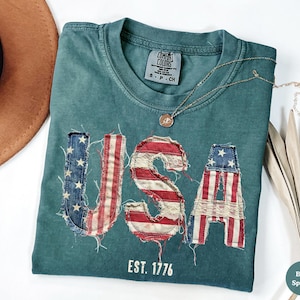 Puede incluir: Una camiseta de color verde azulado con la palabra "USA" en un diseño de bandera estadounidense desgastada. La camiseta tiene el texto "EST. 1776" debajo de las letras. Un collar dorado con un colgante está drapeado sobre la camiseta.