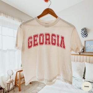 May include: An ivory short-sleeved t-shirt featuring the word GEORGIA printed in a red, distressed, collegiate-style font across the chest. The shirt is displayed on a wooden hanger against a neutral background, showcasing a classic crew neck design.