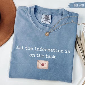 Może przedstawiać: Niebieska koszulka dżinsowa z białym napisem "all the information is on the task". Koszulka ma akwarelową ilustrację koperty. Złoty naszyjnik z wisiorkiem jest zarzucony na koszulkę.