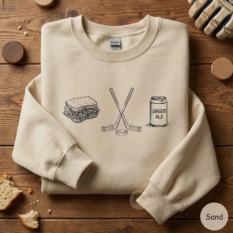 May include: Sand-colored sweatshirt with a black and white graphic of a sandwich, crossed hockey sticks, a hockey puck, and a can of ginger ale. The can is labeled "Ginger Ale".