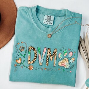 May include: A teal Comfort Colors t-shirt with the letters "DVM" in a watercolor and leopard print design. A stethoscope with a paw print and heart graphic is on the left. A gold necklace with a pendant is on the shirt.