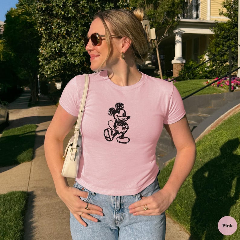 May include: A light pink t-shirt featuring a black sketch of Mickey Mouse. The short-sleeved shirt is paired with blue jeans. The image is taken outdoors on a sunny day.