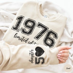 May include: Sand-colored sweatshirt with the year "1976" printed in distressed black lettering. The text "limited edition" is below the numbers. The sleeve features "chapter 55" with a heart graphic. The sweatshirt is folded on a white surface.
