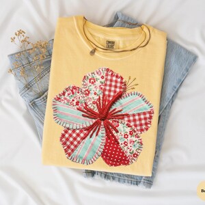 May include: A yellow Comfort Colors t-shirt with a large, appliqued flower design. The flower features red gingham, floral, and polka dot patterns, with a red stitched center. The shirt is folded on top of a pair of blue jeans.