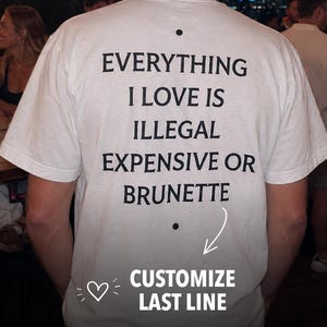 May include: A white short-sleeved t-shirt featuring black text on the back that reads: EVERYTHING I LOVE IS ILLEGAL EXPENSIVE OR BRUNETTE. The design includes a small black dot above and below the text, with a white arrow pointing to the last line.