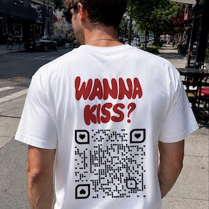 Può includere: Una t-shirt bianca a maniche corte con la scritta rossa "WANNA KISS?" stampata sul retro. Sotto il testo si trova un grande QR code nero composto da minuscole forme a cuore, con un'icona a cuore più grande al centro dei tre quadrati d'angolo.
