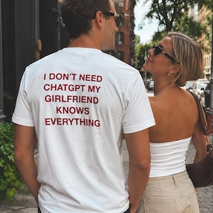 May include: A white short-sleeved t-shirt featuring dark red text on the back that reads: I DON'T NEED CHATGPT MY GIRLFRIEND KNOWS EVERYTHING. The shirt is made of a plain fabric and has a standard crew neckline, shown from a rear-facing perspective.