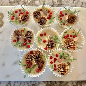 May include: Nine festive, cream-colored wax melts in paper cups, each decorated with pinecones, red berries, and sprigs of greenery. The arrangement sits on a white marble tray with a gold letter "B".
