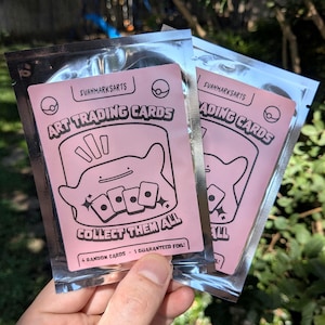 Peut inclure: Deux paquets en aluminium argenté contenant des cartes à collectionner. Les étiquettes roses portent le texte EVANMARKSARTS, ART TRADING CARDS, COLLECT THEM ALL, et 4 RANDOM CARDS - 1 GUARANTEED FOIL, avec un dessin au trait d'une créature.
