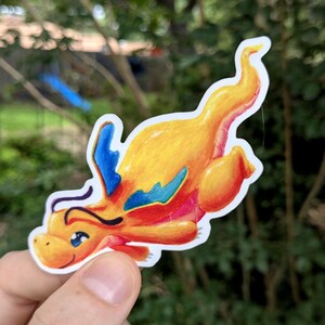 May include: A die-cut sticker featuring a stylized orange dragon with a long, curved tail and small blue wings. The dragon has a rounded head, a small smile, and large blue eyes. The illustration uses vibrant orange, yellow, and red tones.