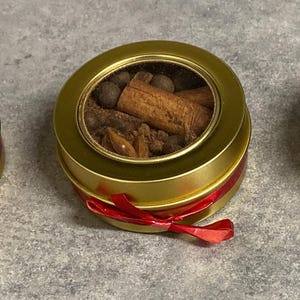 May include: A gold-coloured round tin filled with various spices, including cinnamon sticks and peppercorns, visible through a clear lid. A red ribbon is tied around the tin, forming a bow.