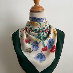 May include: A square silk scarf featuring a cream-colored center with a vibrant floral and butterfly print in shades of blue, red, and yellow. The scarf has a wide, solid dark green border and includes the Gucci brand name printed on the fabric.